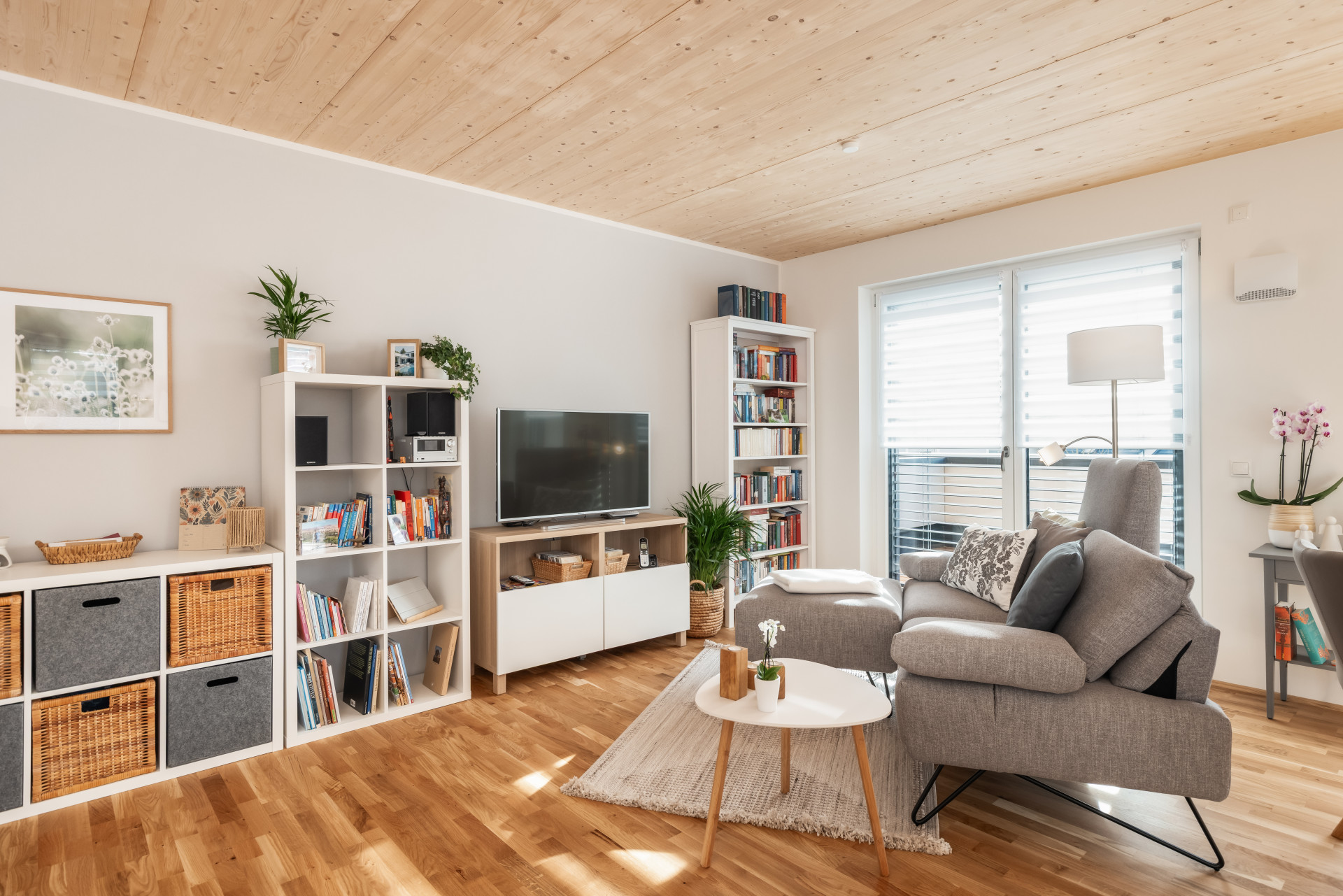 Modernes Wohnzimmer mit grauem Sofa, Regalwand, Holzdecke, Fernseher, runden Couchtisch und großer Fensterfront mit Jalousien.