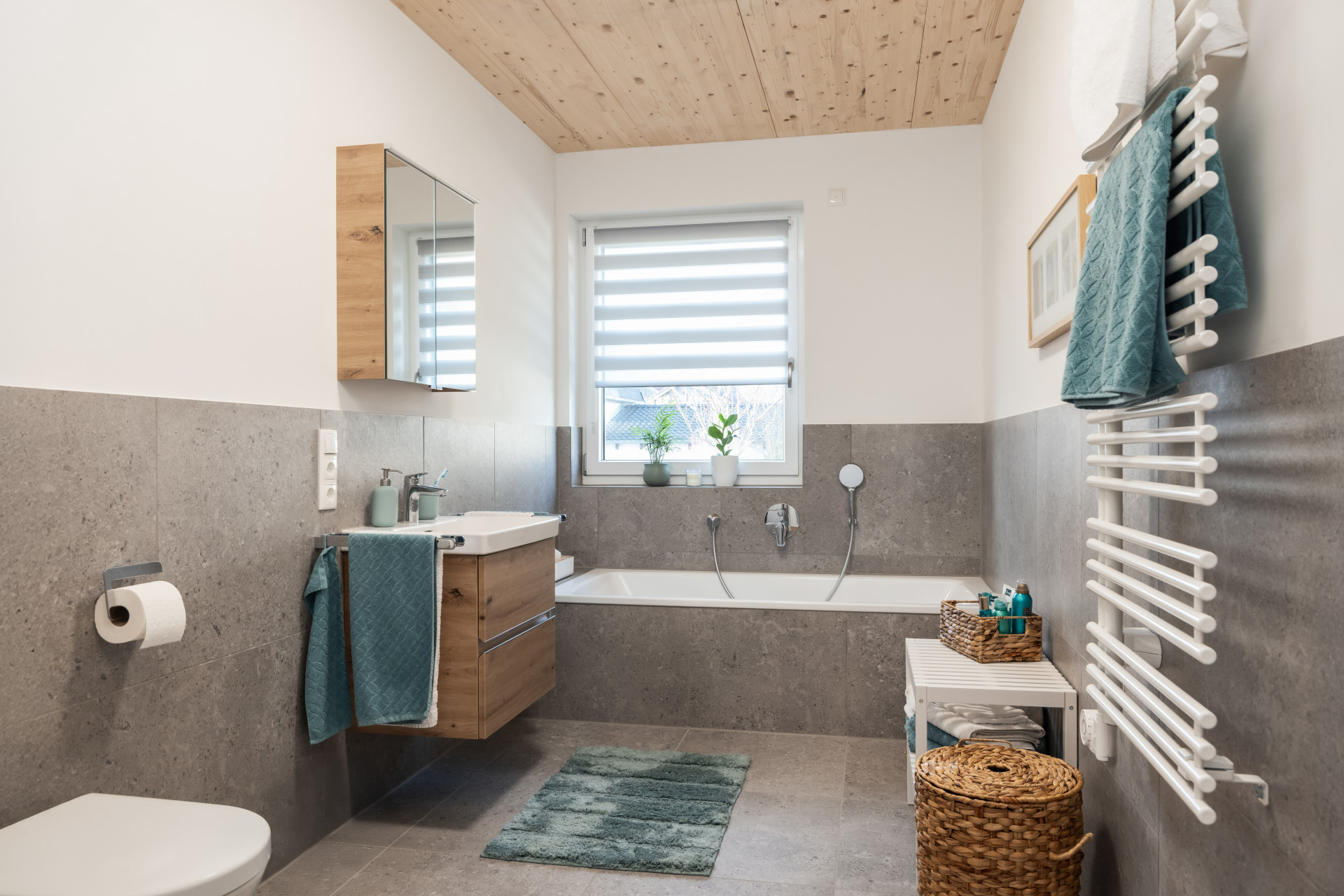 Modernes Badezimmer mit Badewanne, Holzdecke, grauen Fliesen, weißen Handtuchhaltern, Pflanzen, und türkisen Handtüchern.