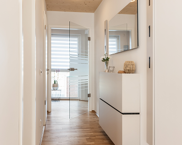 Ein heller Flur mit Holzfußboden, einem Spiegel an der Wand und einem Glasfenster. Eine Kommode mit Dekoration steht daneben.